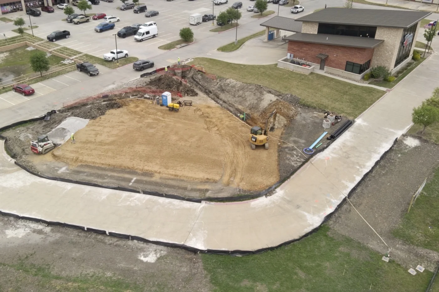 Aerial view of the Anna site in April 2026, showing the excavated building pad, the newly poured access drive curving around it, and the neighbouring buildings and road beyond.