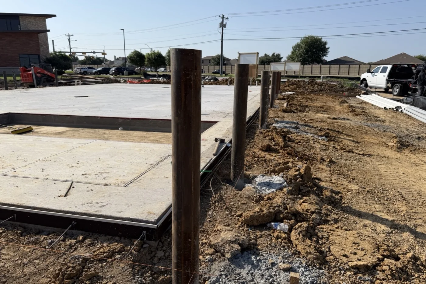The poured building slab at the Anna site in July 2026, with a row of steel columns standing in their footings along its edge.
