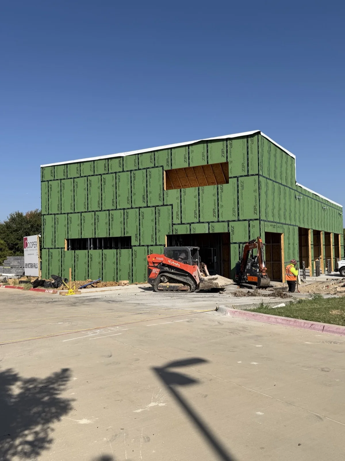 The Anna, Texas facility in September 2026: the building wrapped in green sheathing, a row of open service-bay openings along its right-hand side, construction equipment working at the base and the poured parking lot in the foreground.