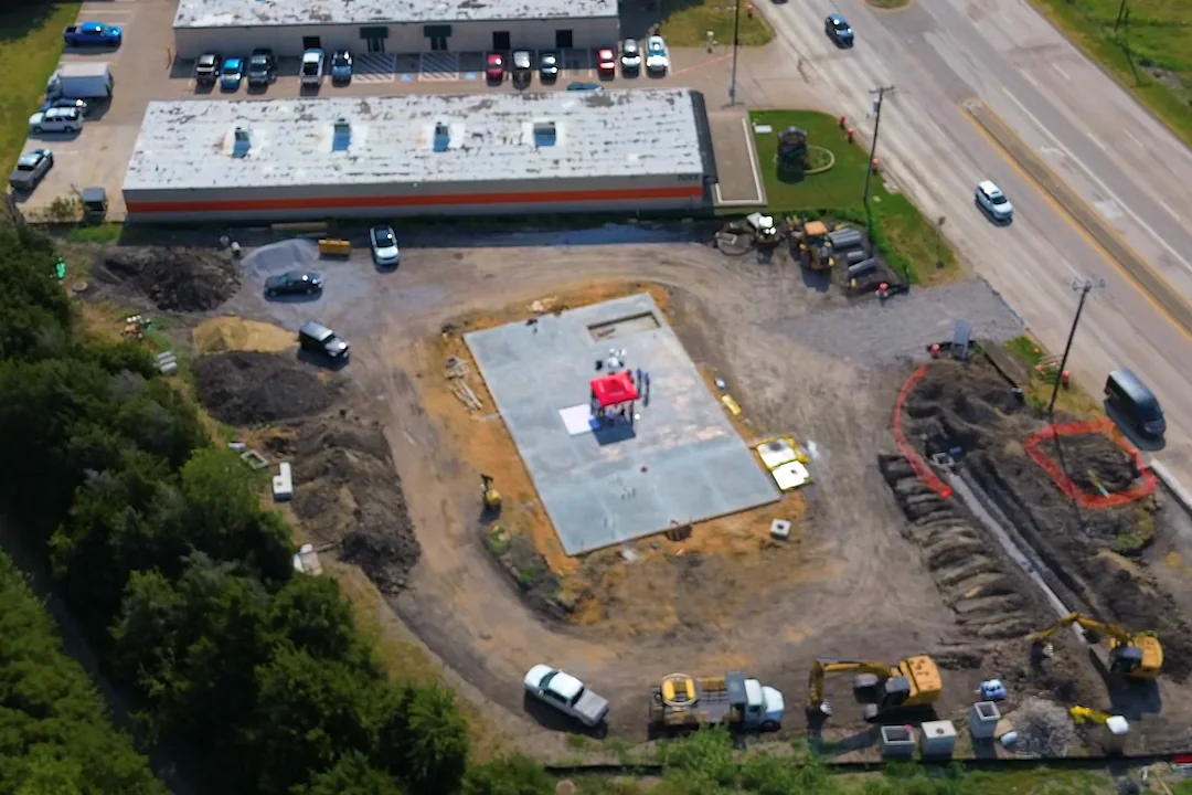 Aerial view of the Wylie development site during construction. State Highway 78 runs down the right-hand edge; the poured building slab sits at the centre of the parcel with open utility trenching around it; the graded vehicle approach lies between the slab and the highway; and open ground for the service side and future expansion runs along the left.