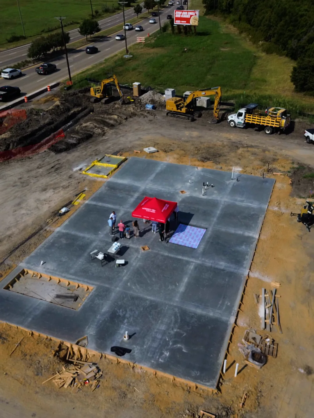 Aerial view looking down on the poured concrete slab at the Wylie, Texas site, with formwork and plumbing stub-ups across it and excavators working at its edge.